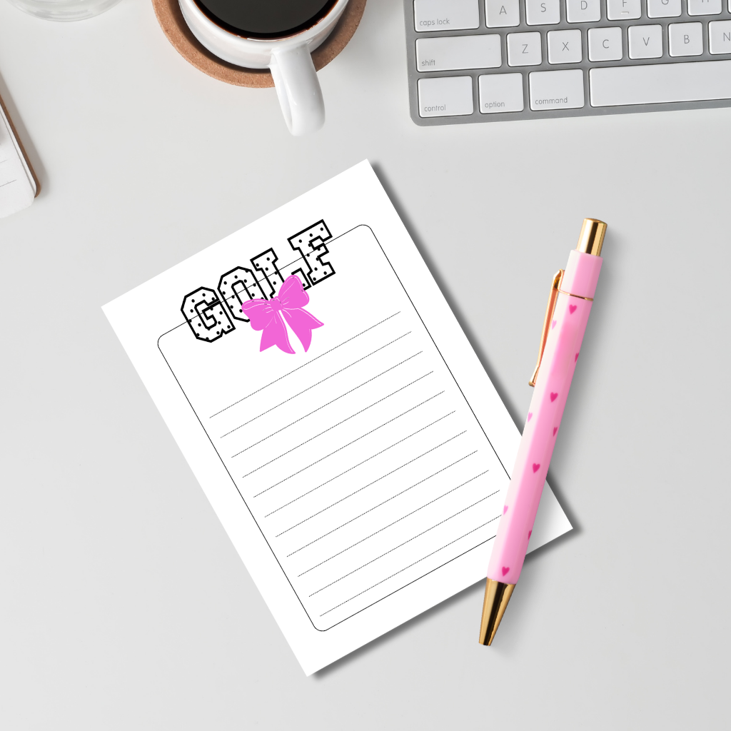 A styled flat-lay of a white lined notepad with the word 'GOLF' and a pink bow graphic, paired with a pink pen that has small heart details and gold accents, on a light gray desk with a keyboard, coffee cup, and notebook partially visible.