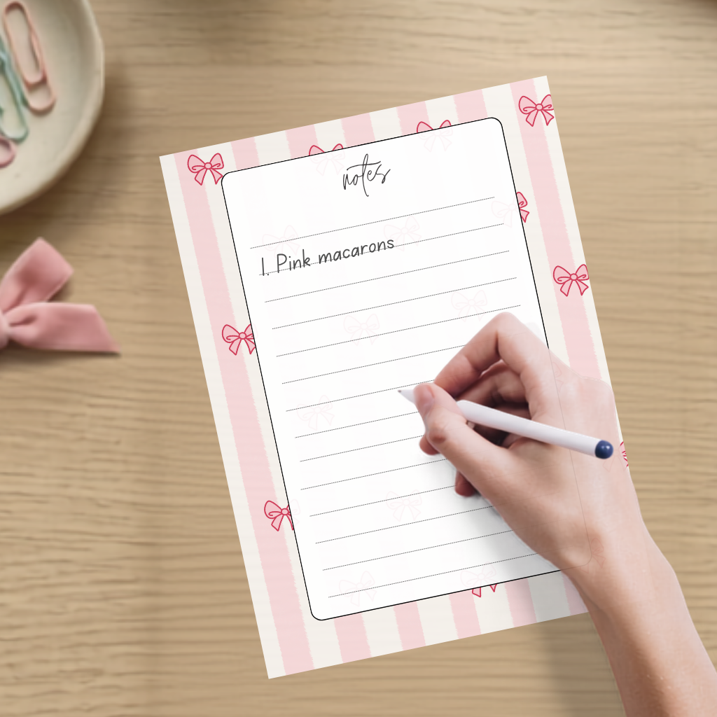 A hand holding a white pen writes on a lined notepad with pink and white stripes and small bow illustrations. The notepad is placed on a light wood desk, with a pink ribbon and pastel paperclips visible in the background.