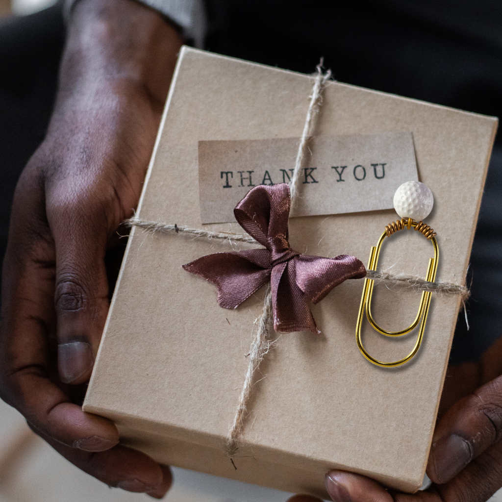 A gold paperclip with a white golf ball bead attached with gold colored wire, shown on a square kraft gift box with twine and a shiny brown bow, with a paper tag that reads 'Thank You' being held in a person's hand.