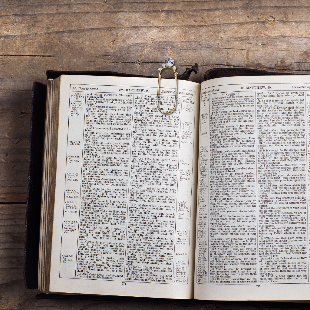 An open vintage dictionary or book with dense printed text on both pages, held together by a gold-toned metal bookmark shaped like a rose with blue gemstone accents, placed on a rustic wooden surface.