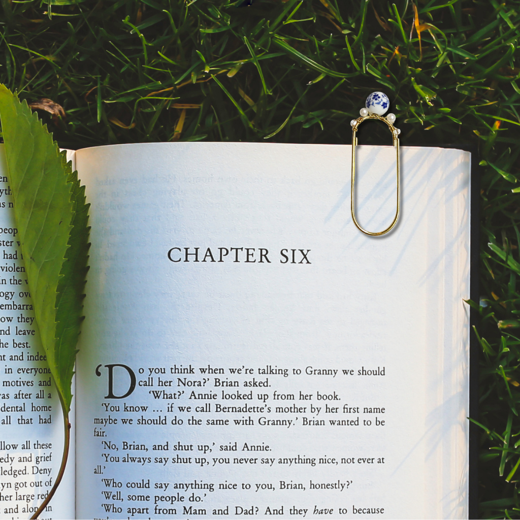A gold metal paperclip bookmark with a blue and white floral bead holding an open book outdoors on green grass.
