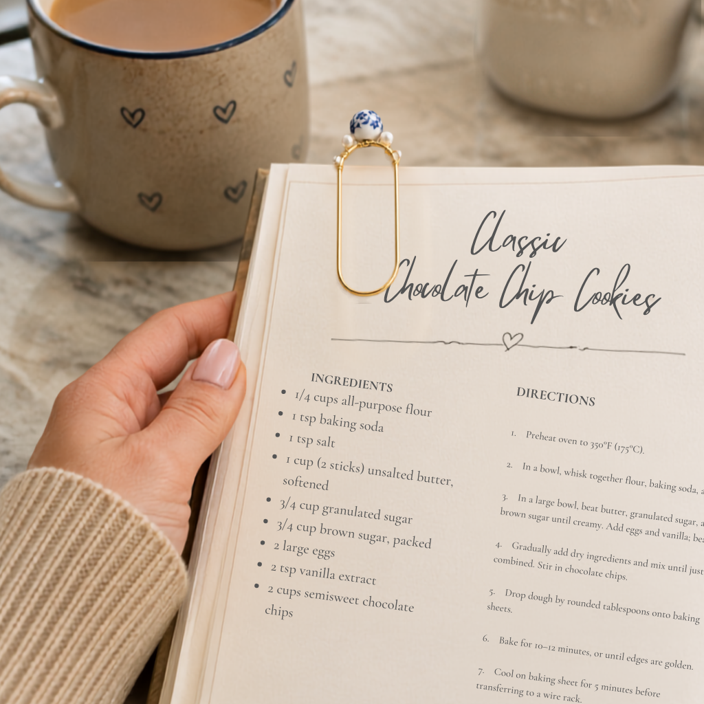 A hand holds an open recipe book page titled 'Classic Chocolate Chip Cookies' with ingredients and directions visible. A gold paperclip with a blue and white bead and smaller white beads clipped onto the page, and a ceramic mug is blurred in the background.