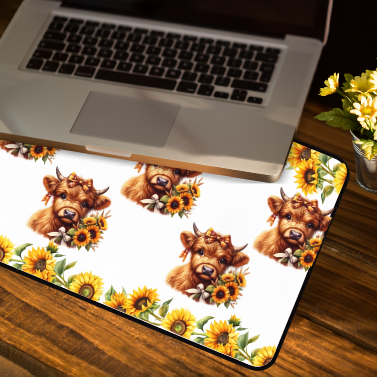 A rectangular desk mat with a white background and a repeating print of Highland cows holding sunflowers, styled on a wooden desk next to an open laptop and a vase of yellow flowers.