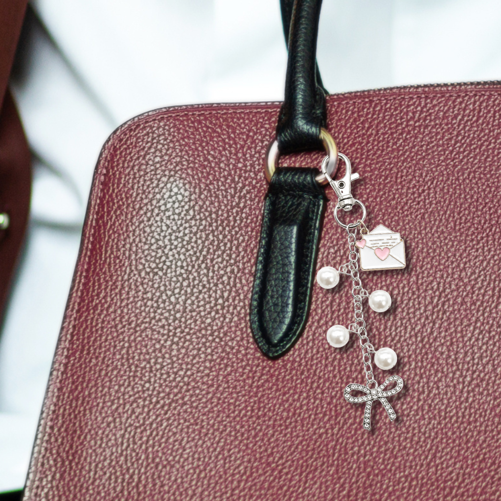 A close-up of a burgundy pebbled leather handbag with a black leather handle, adorned with a silver-tone charm featuring a heart-shaped envelope, pearls, and a bow.