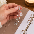 A hand with light pink manicured nails holds a silver-toned charm keychain featuring pearl beads, a bow charm, and a small envelope charm with a pink heart, set against a background of a brown planner cover and an open notebook.