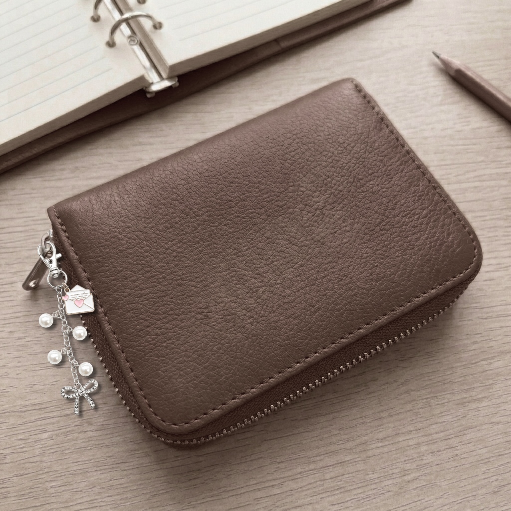 A brown textured leather zip-around card holder wallet with silver-tone zipper hardware and dangling charms, shown on a light wood surface with a notebook and pencil in the background.