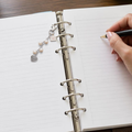 A silver charm keychain with pearls and a filagree heart pendant rests on an open lined ring binder notebook. A hand holding a pen is visible on the right side, and the notebook lies on a dark wooden surface.