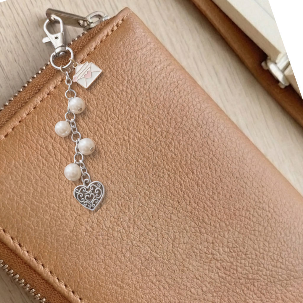 A close-up of a tan textured leather wallet or notebook cover with a silver-tone zipper pull charm featuring white pearl beads, a small envelope charm, and a heart-shaped filigree ornament.