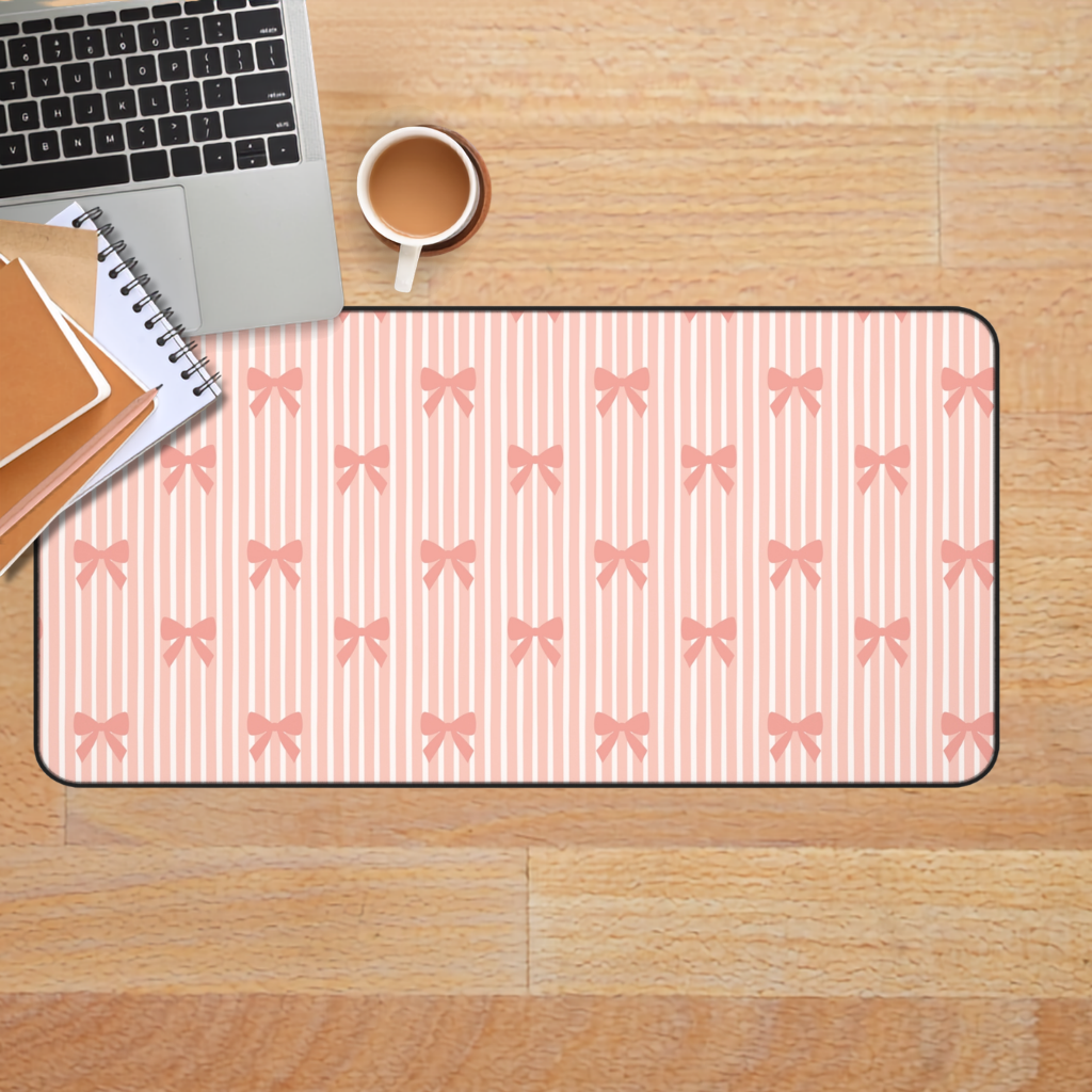 A rectangular desk mat with a pink and white striped pattern and repeating bow motifs, styled on a wooden desk next to a laptop, notebook, and coffee cup.