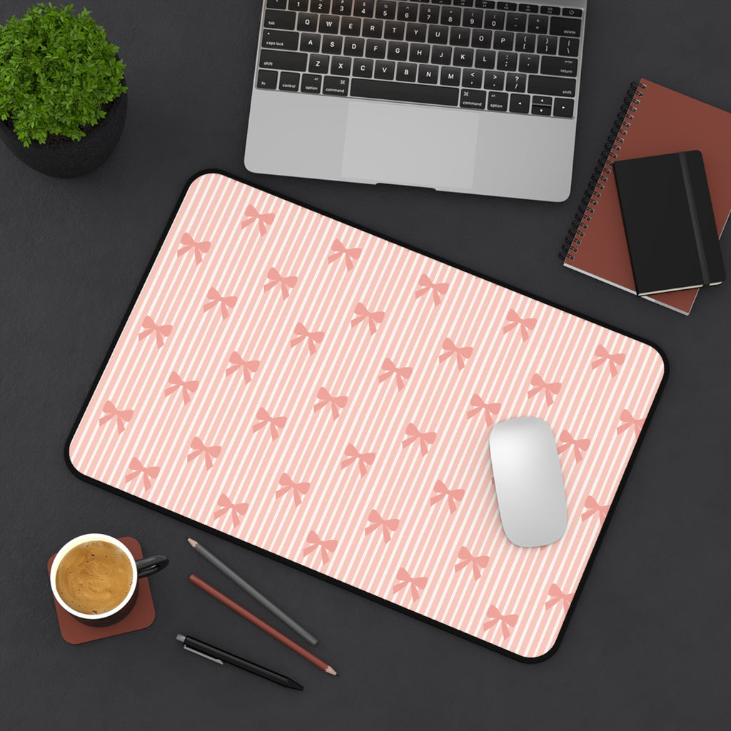 A large desk mat with a pink and white striped pattern and a repeating bow motif, styled in a flat-lay workspace scene with a laptop, mouse, coffee cup, and notebooks on a dark gray surface.