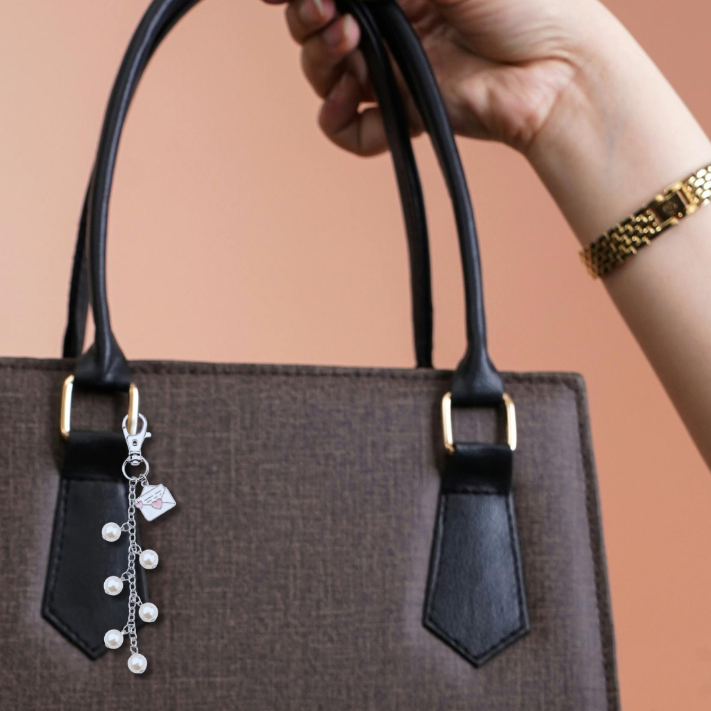 A close-up of a brown textured handbag with black leather handles and gold-tone hardware, featuring a dangling silver charm with pearl-like beads and a small envelope charm with a pink heart. A hand holding the bag is visible against a solid peach background.