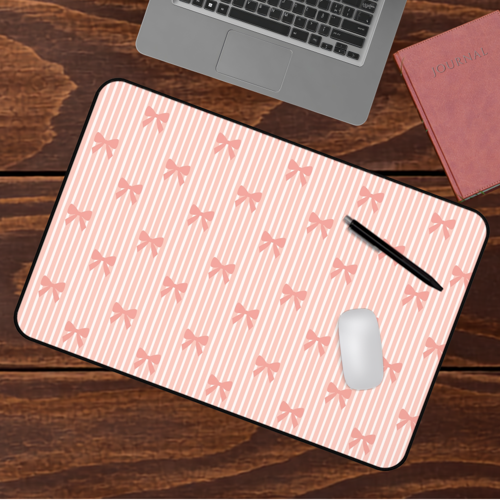 A rectangular desk mat with a pink and white striped pattern decorated with small pink bows, shown on a dark wood desk with a laptop, mouse, pen, and notebook nearby.